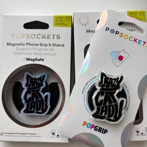 May include: Two PopSockets phone grips and stands. One is packaged in a white box with the text "POPSOCKETS" and "CRAZY CAT LADY" in a black and white design. The other is on a white backing with the same design.