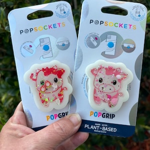 May include: Two PopSockets phone grips featuring a cow design with strawberry accents. The grips are white with a clear, glittery top. The packaging reads "POPSOCKETS" and "POPGRIP" with "made with PLANT-BASED materials".
