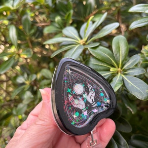 May include: Black heart-shaped resin charm with a white design of two hands reaching up towards a moon and stars. The charm is filled with iridescent glitter and attached to a clear retractable badge holder.