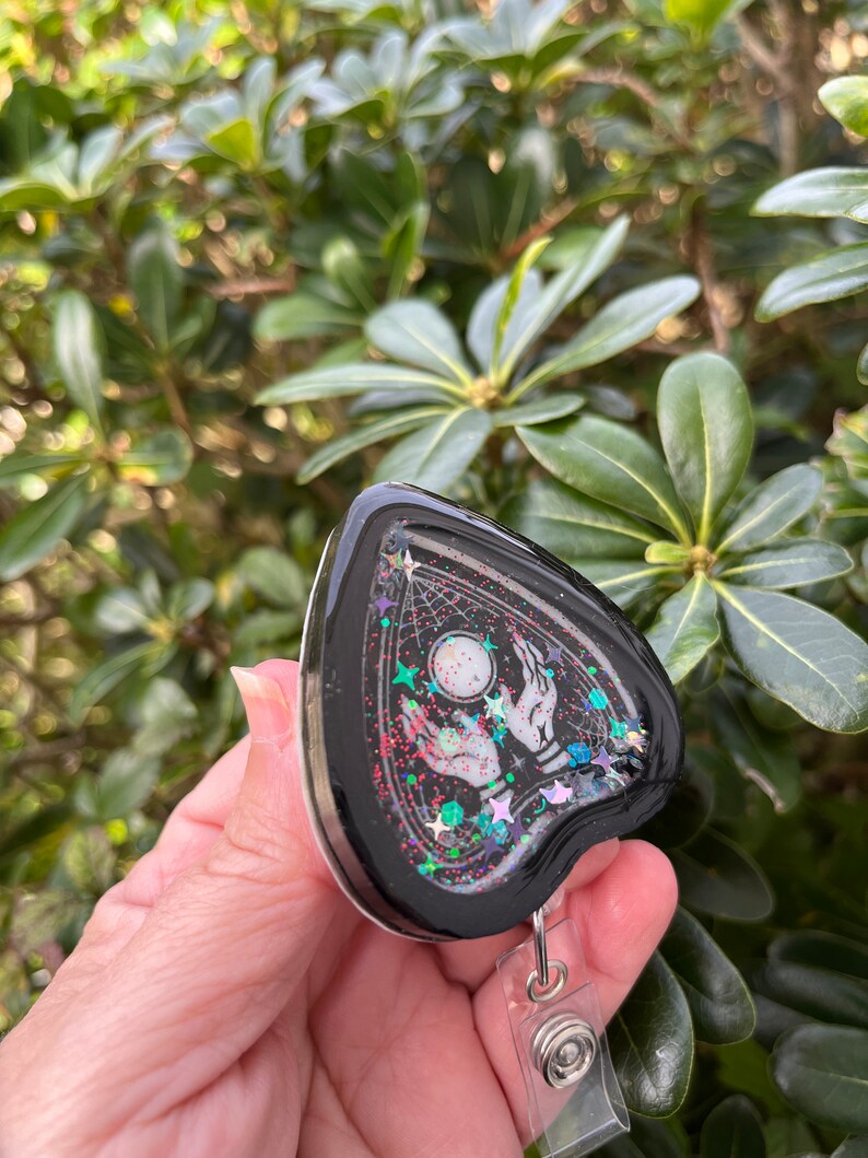 May include: Black heart-shaped resin charm with a white design of two hands reaching up towards a moon and stars. The charm is filled with iridescent glitter and attached to a clear retractable badge holder.