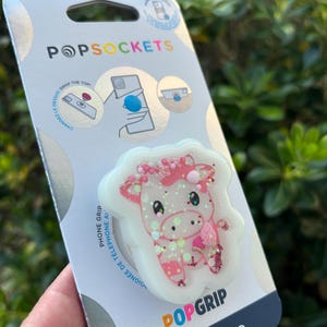 May include: A PopSockets PopGrip phone grip featuring a pink and white cow design with glitter accents. The packaging includes the PopSockets logo and the text "made with PLANT-BASED materials."