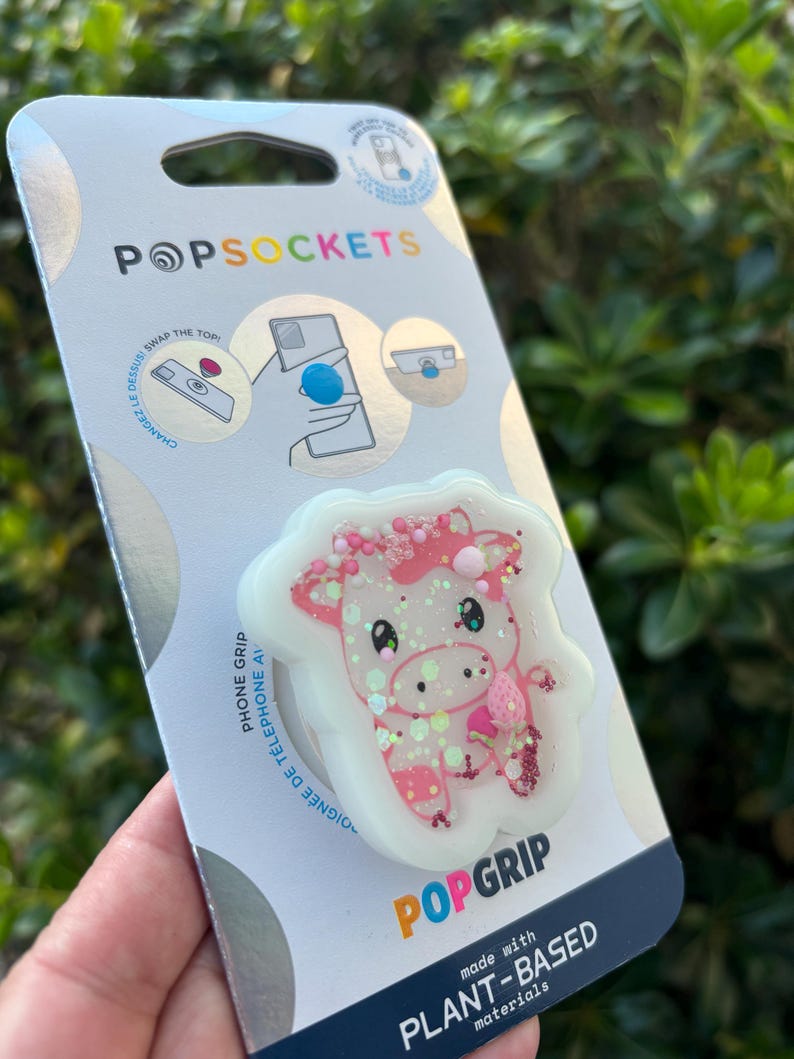 May include: A PopSockets PopGrip phone grip featuring a pink and white cow design with glitter accents. The packaging includes the PopSockets logo and the text "made with PLANT-BASED materials."