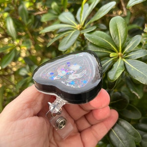 May include: A black heart-shaped resin charm with a pentagram design, glitter, and a silver-toned charm. The charm is attached to a clear plastic retractable badge holder.