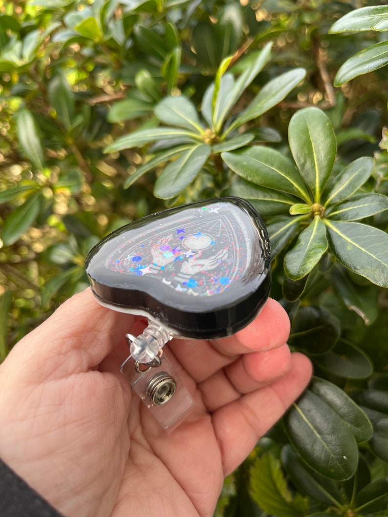 May include: A black heart-shaped resin charm with a pentagram design, glitter, and a silver-toned charm. The charm is attached to a clear plastic retractable badge holder.