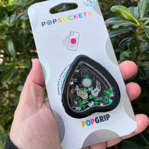 Pu&ograve; includere: Un PopGrip per telefono nero e verde con un design glitterato. Il design presenta un cuore nero con un contorno bianco, una sfera di cristallo e due mani che raggiungono la sfera. Il testo "POPGRIP" &egrave; stampato in bianco sul fondo della presa.