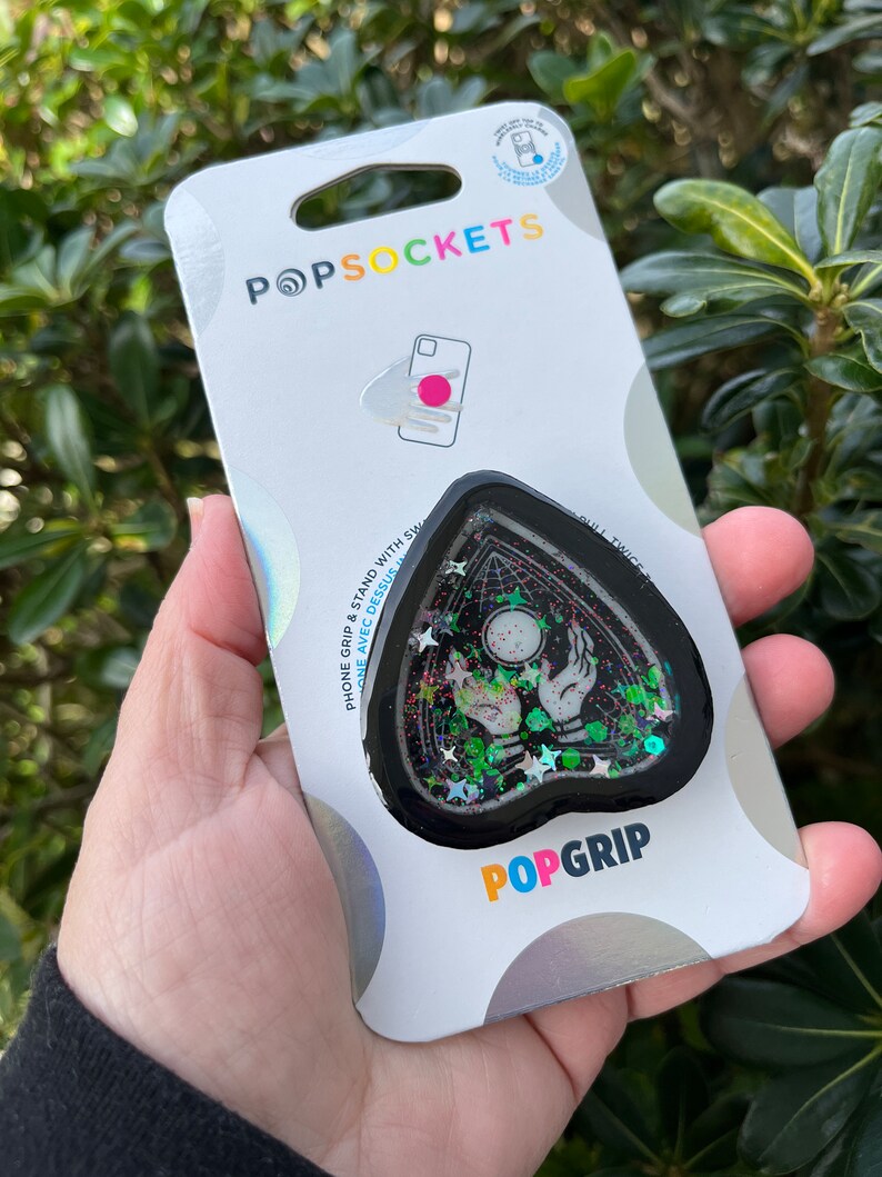 Pu&ograve; includere: Un PopGrip per telefono nero e verde con un design glitterato. Il design presenta un cuore nero con un contorno bianco, una sfera di cristallo e due mani che raggiungono la sfera. Il testo "POPGRIP" &egrave; stampato in bianco sul fondo della presa.