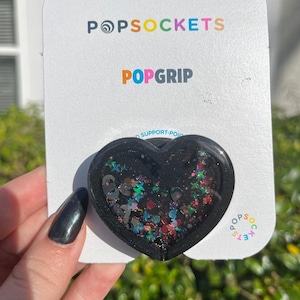 May include: Black heart-shaped PopGrip phone grip with iridescent glitter and stars. The PopGrip is on a white card with the PopSockets logo and the text "PopGrip".