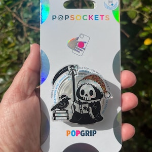 May include: A white and black PopGrip phone grip featuring a cartoon Grim Reaper wearing a Santa hat and holding a book. A black crow with a Santa hat is perched on a stack of books. The text "PHONE GRIP" and "ELEPHONE AVEC DESSUS" is printed on the grip. The text "POPGRIP" is printed on the packaging.