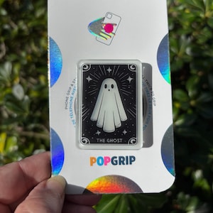 May include: A PopGrip phone grip featuring a white ghost illustration on a black background with the text "THE GHOST." The grip is rectangular with rounded corners and is packaged on a white and iridescent card.