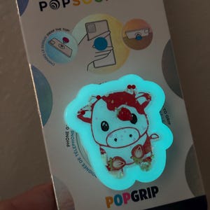 May include: A PopSockets PopGrip featuring a glowing cow design with strawberries. The PopGrip is white with red accents and a light blue outline. The packaging includes the text "POPSOCKETS" and "POPGRIP", and states it is made with plant-based materials.