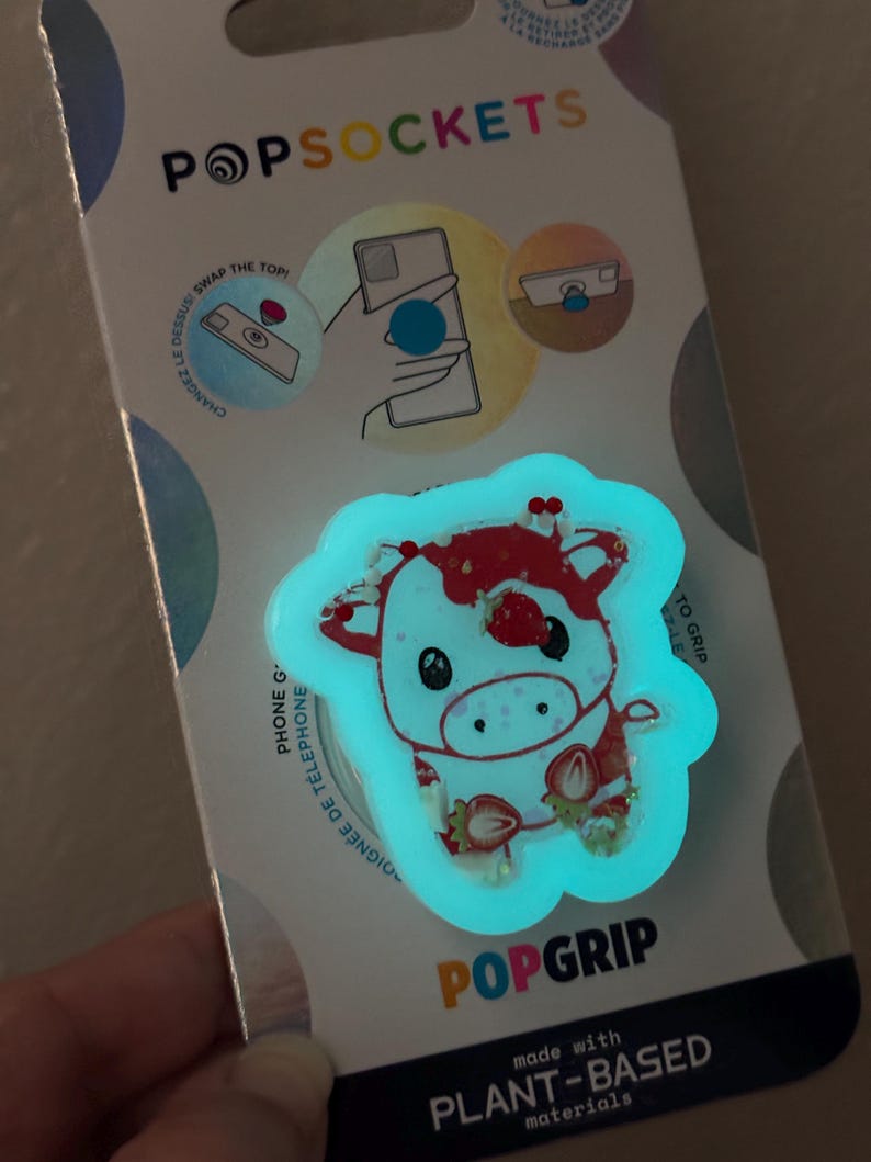 May include: A PopSockets PopGrip featuring a glowing cow design with strawberries. The PopGrip is white with red accents and a light blue outline. The packaging includes the text "POPSOCKETS" and "POPGRIP", and states it is made with plant-based materials.