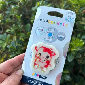 May include: A PopSockets PopGrip featuring a cow design with red and white accents, and strawberry details. The PopGrip is on a white backing with the PopSockets and PopGrip logos. The packaging states "made with plant-based materials."