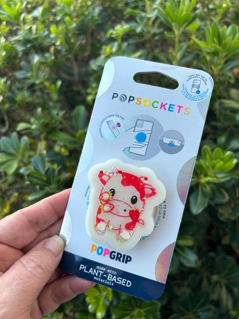 May include: A PopSockets PopGrip featuring a cow design with red and white accents, and strawberry details. The PopGrip is on a white backing with the PopSockets and PopGrip logos. The packaging states "made with plant-based materials."
