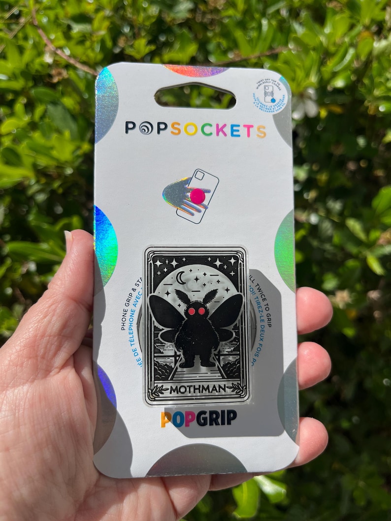 Puede incluir: Una agarradera de tel&eacute;fono blanca y plateada con una ilustraci&oacute;n en blanco y negro de un mothman. La agarradera dice "Mothman" y "PopGrip".