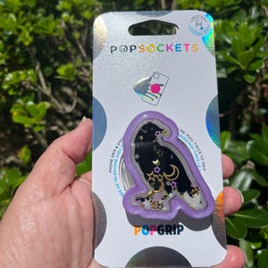 May include: A PopSockets PopGrip featuring a purple owl design with a black interior filled with gold stars, moons, and glitter. The PopGrip is attached to a silver backing with the PopSockets and PopGrip logos.