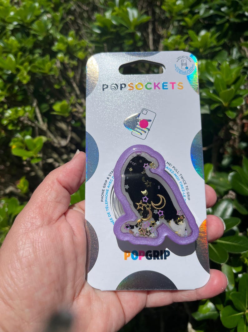 May include: A PopSockets PopGrip featuring a purple owl design with a black interior filled with gold stars, moons, and glitter. The PopGrip is attached to a silver backing with the PopSockets and PopGrip logos.