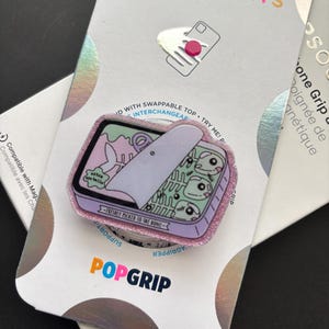 May include: A phone grip featuring a rectangular tin illustration with a light purple border and glittery edges. The graphic shows a partially opened lid revealing fish skeletons with large eyes, a fish bone, and the text "extra eye balls" and "FRESHLY PICKED TO THE BONE".