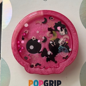 May include: A pink PopGrip with a skull design. The skull is black and has a cartoonish appearance. The PopGrip is filled with pink liquid and decorative elements, including stars, moons, and other small shapes. The word "POPGRI" is visible.