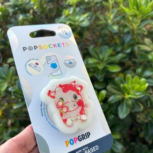 May include: A PopSockets PopGrip phone grip featuring a red and white cow design with strawberry accents. The grip is made with plant-based materials and is attached to a white backing with the PopSockets logo.
