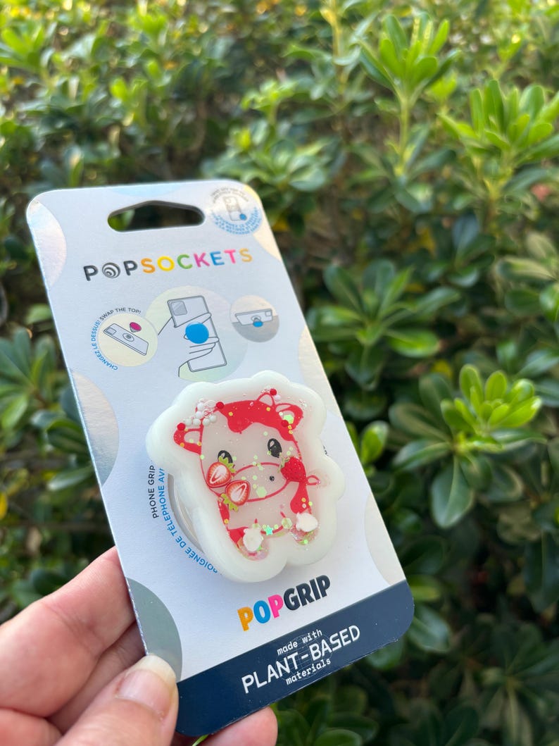 May include: A PopSockets PopGrip phone grip featuring a red and white cow design with strawberry accents. The grip is made with plant-based materials and is attached to a white backing with the PopSockets logo.