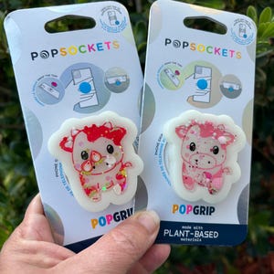 May include: Two PopSockets phone grips featuring cow designs. One is red and the other is pink, both with glitter accents. The packaging includes the PopSockets logo and the words "POPGRIP" and "PLANT-BASED materials".