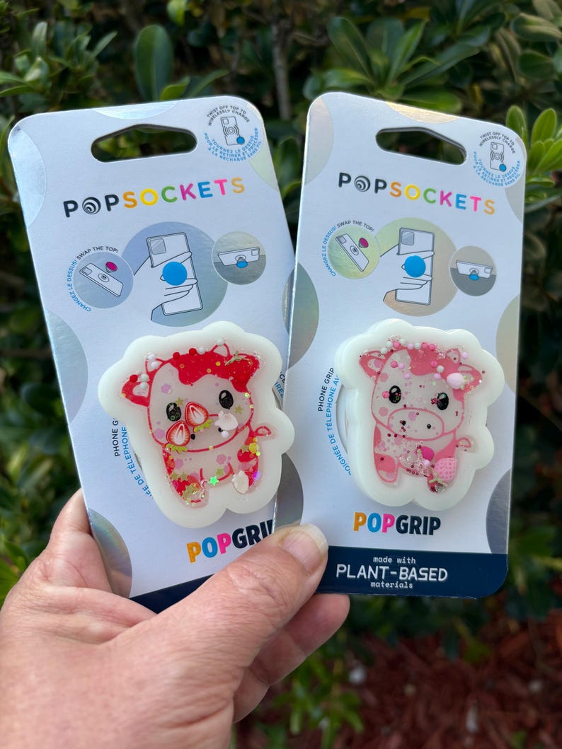 May include: Two PopSockets phone grips featuring cow designs. One is red and the other is pink, both with glitter accents. The packaging includes the PopSockets logo and the words "POPGRIP" and "PLANT-BASED materials".