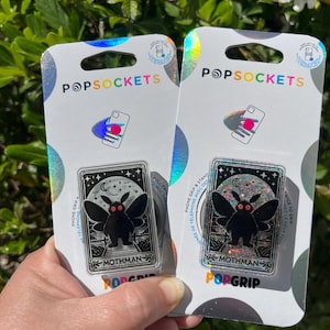 Puede incluir: Dos agarres para tel&eacute;fono PopSockets con una ilustraci&oacute;n en blanco y negro de un mothman con ojos rojos. El texto "Mothman" est&aacute; impreso debajo de la ilustraci&oacute;n. El fondo de la ilustraci&oacute;n es negro con estrellas blancas y una luna creciente.