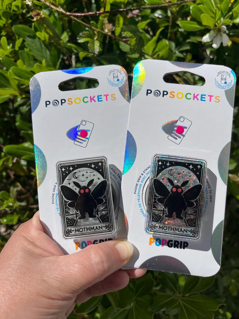 Puede incluir: Dos agarres para tel&eacute;fono PopSockets con una ilustraci&oacute;n en blanco y negro de un mothman con ojos rojos. El texto "Mothman" est&aacute; impreso debajo de la ilustraci&oacute;n. El fondo de la ilustraci&oacute;n es negro con estrellas blancas y una luna creciente.