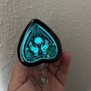 May include: A black heart-shaped resin charm with a glow-in-the-dark design of a Ouija board with hands reaching up to a glowing moon. The charm is attached to a clear plastic retractable badge holder.