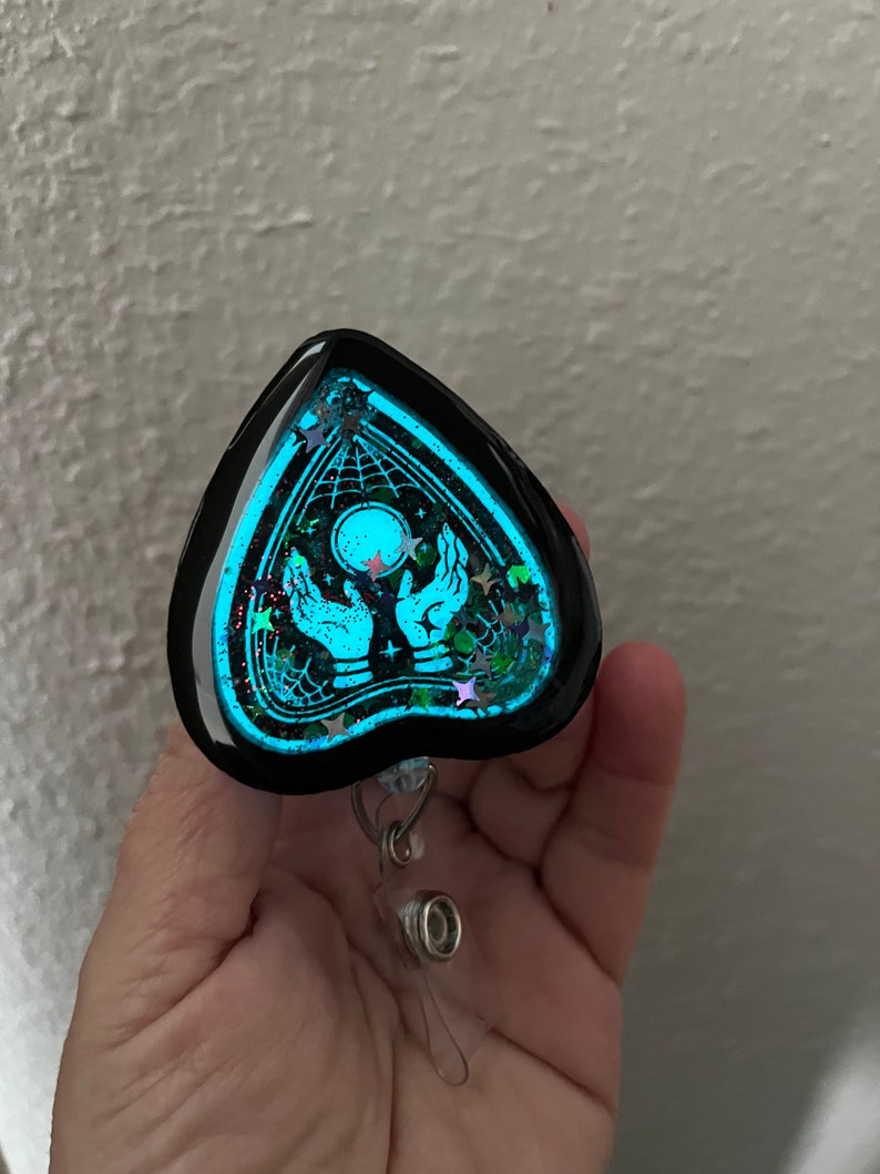 May include: A black heart-shaped resin charm with a glow-in-the-dark design of a Ouija board with hands reaching up to a glowing moon. The charm is attached to a clear plastic retractable badge holder.