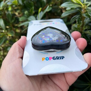Pu&ograve; includere: Un'impugnatura per telefono a forma di cuore nero con un design scintillante che presenta una luna crescente, stelle e una mano. L'impugnatura &egrave; confezionata su una carta bianca con il testo "POPGRIP" e "Grip/Support".