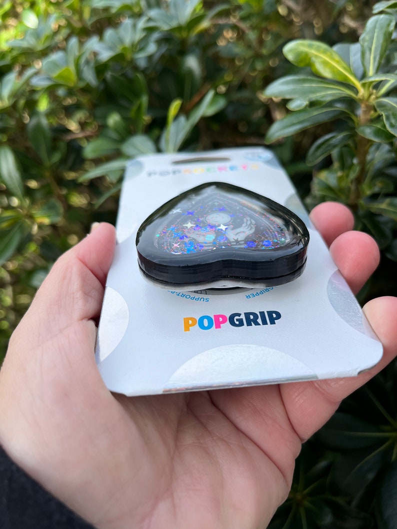 Pu&ograve; includere: Un'impugnatura per telefono a forma di cuore nero con un design scintillante che presenta una luna crescente, stelle e una mano. L'impugnatura &egrave; confezionata su una carta bianca con il testo "POPGRIP" e "Grip/Support".