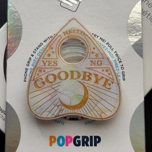 May include: A triangular planchette-shaped phone grip with an iridescent finish and gold-colored text reading HELLO, YES, NO, and GOODBYE. The design features a crescent moon and star accents. It is mounted on a white backing card labeled POPGRIP.