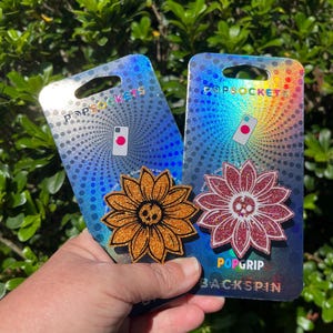 May include: Two PopSockets PopGrip Backspin phone accessories. One is orange glitter with a sunflower design and a skull center. The other is red glitter with a sunflower design and a skull center. Both are on holographic backing.