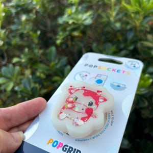 May include: A white PopSocket with a red and white cow design, featuring strawberry accents and glitter. The PopSocket is attached to a white and blue branded backing that reads "PopGrip" and "made with plant-based materials."
