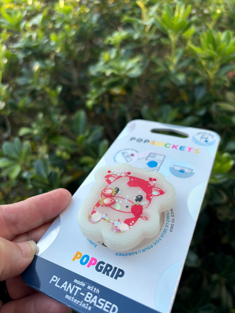 May include: A white PopSocket with a red and white cow design, featuring strawberry accents and glitter. The PopSocket is attached to a white and blue branded backing that reads "PopGrip" and "made with plant-based materials."