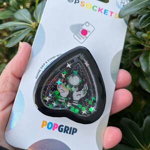 Pu&ograve; includere: Un'impugnatura per telefono nera a forma di cuore con un design scintillante. Il design presenta una sfera di cristallo, due mani e stelle. L'impugnatura &egrave; confezionata su una carta bianca con il testo "PopSockets" e "PopGrip".