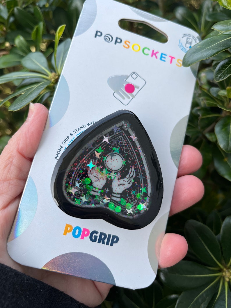 Pu&ograve; includere: Un'impugnatura per telefono nera a forma di cuore con un design scintillante. Il design presenta una sfera di cristallo, due mani e stelle. L'impugnatura &egrave; confezionata su una carta bianca con il testo "PopSockets" e "PopGrip".