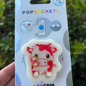 May include: A PopSockets PopGrip with a red and white cow design, holding strawberries. The PopGrip is made with plant-based materials and is attached to a card with the PopSockets and PopGrip logos.