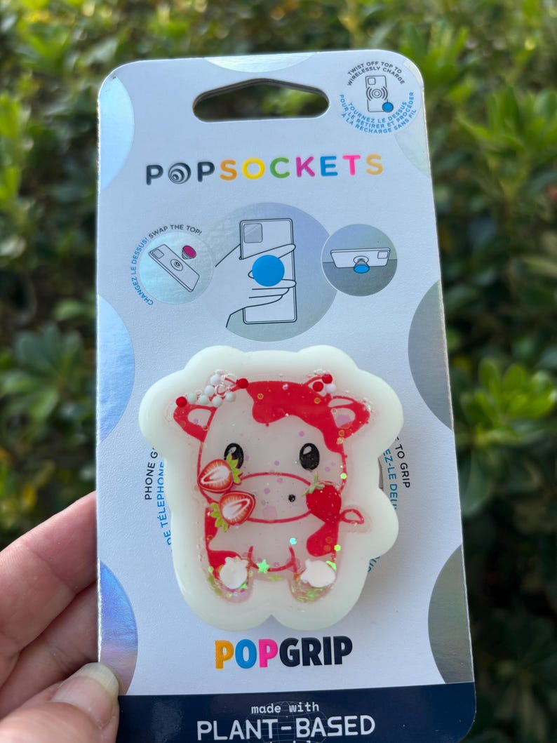 May include: A PopSockets PopGrip with a red and white cow design, holding strawberries. The PopGrip is made with plant-based materials and is attached to a card with the PopSockets and PopGrip logos.