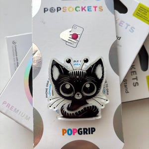 May include: A PopSockets PopGrip phone accessory featuring a black and white illustration of a stylized cat with large eyes and antennae. The product is mounted on its original white retail packaging with the text PopSockets and PopGrip visible.