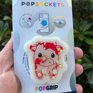 May include: A PopSockets PopGrip featuring a red and white cow design with strawberry accents. The grip is made with plant-based materials and has the text "POPGRIP" and "POPSOCKETS". The packaging includes instructions and the text "SWAP THE TOP!"