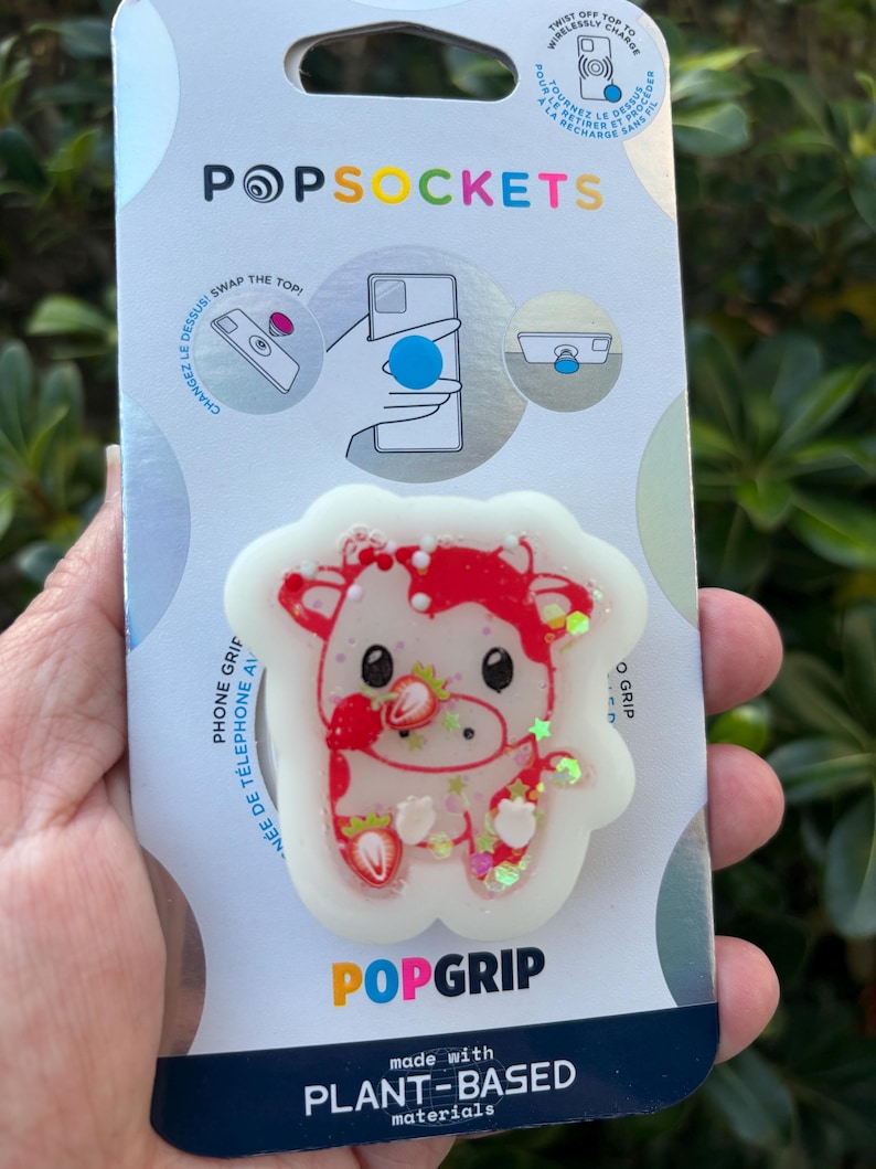 May include: A PopSockets PopGrip featuring a red and white cow design with strawberry accents. The grip is made with plant-based materials and has the text "POPGRIP" and "POPSOCKETS". The packaging includes instructions and the text "SWAP THE TOP!"