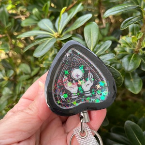May include: Black triangle-shaped keychain with a clear resin insert featuring a white hand holding a glowing orb, surrounded by glitter and stars. The keychain has a silver retractable cord and a silver ring.
