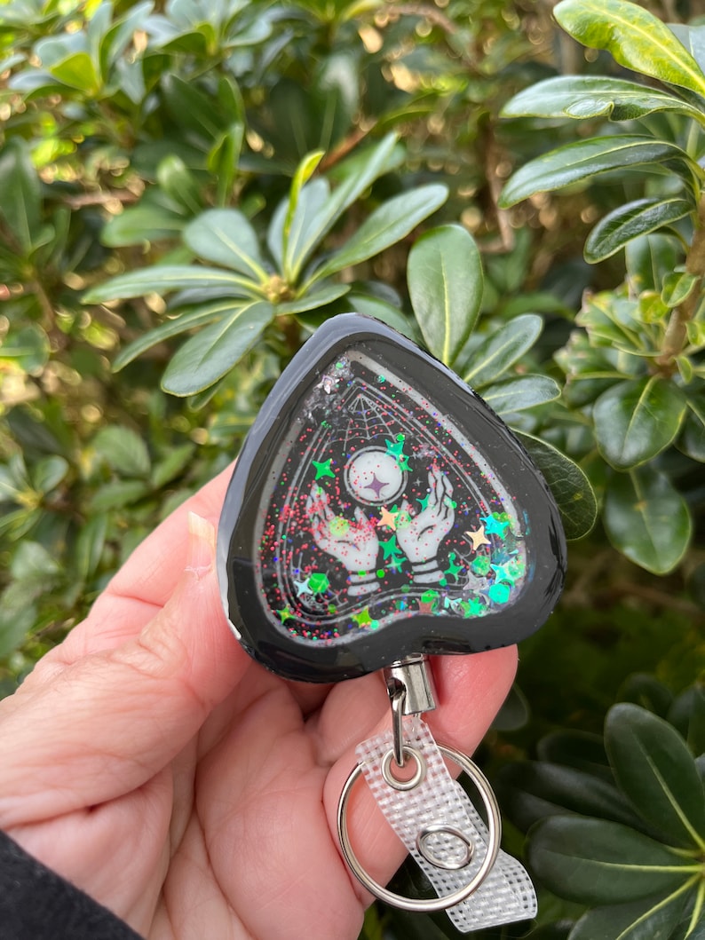 May include: Black triangle-shaped keychain with a clear resin insert featuring a white hand holding a glowing orb, surrounded by glitter and stars. The keychain has a silver retractable cord and a silver ring.