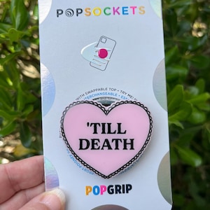 May include: Pink heart-shaped phone grip with a black border and the text 'TILL DEATH' in black lettering. The grip is attached to a white card with the word 'POPGRIP' in pink, blue, and yellow letters.
