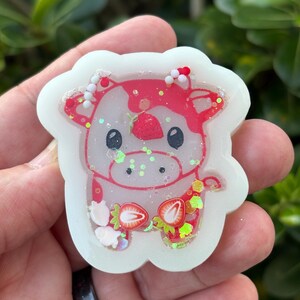 May include: A white, strawberry-themed cow-shaped charm. The cow has a red and pink design with strawberry accents and glitter. The charm is held in a hand against a blurred green background.