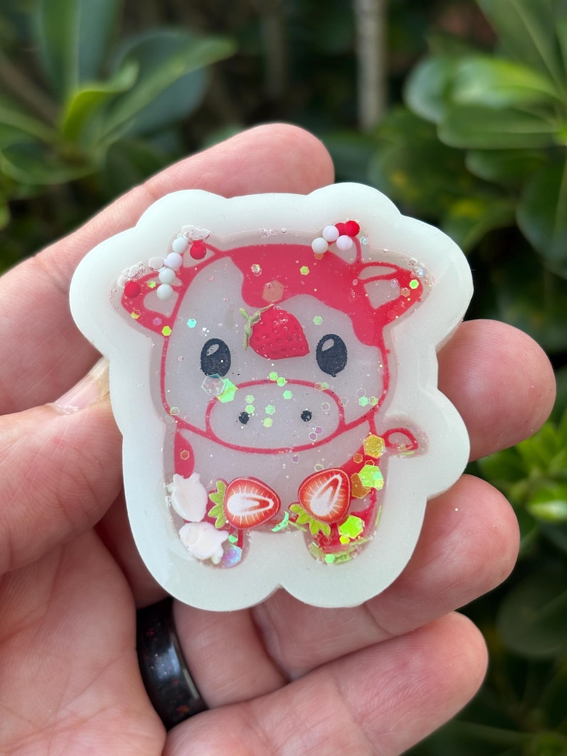 May include: A white, strawberry-themed cow-shaped charm. The cow has a red and pink design with strawberry accents and glitter. The charm is held in a hand against a blurred green background.