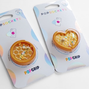 May include: Two PopSockets phone grips with a honeycomb and bee design. One grip is round and the other is heart-shaped. Both grips are yellow and feature a bee, honeycomb, and flowers.
