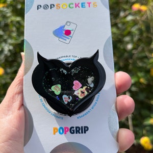 May include: Black phone grip with a heart-shaped design filled with glitter and colorful candy hearts. The text "SUCK YOU" and "BIY ME" are visible on the candy hearts. The phone grip is packaged on a white card with the text "POPSOCKETS" and "POPGRIP".